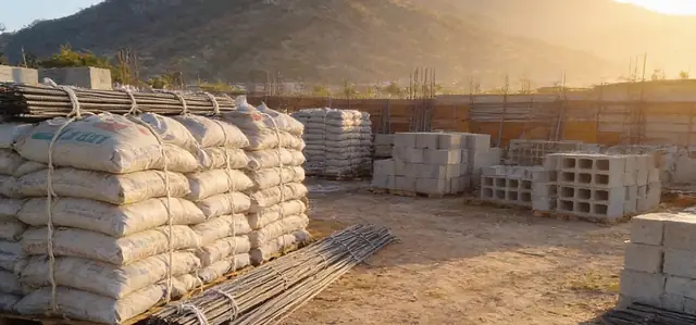 Bultos de cemento, bloques de concreto y varillas de hierro apilados en un patio de materiales, con la montaña al fondo